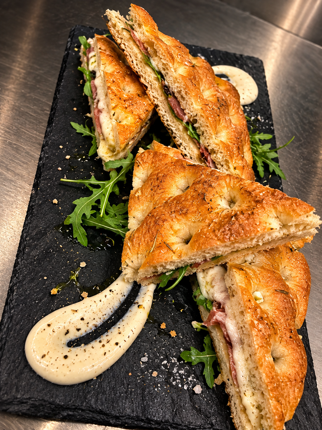 Toast di focaccia farcito con affettati, formaggio filante e rucola, servito su ardesia